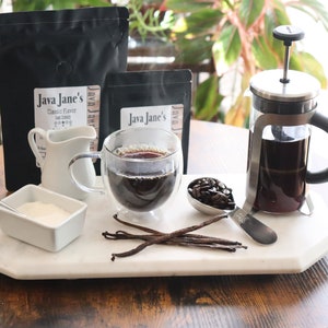 May include: A coffee setup featuring a French press, a double-walled glass mug with coffee, and a small creamer. Also includes a small dish of sugar, a spoon, coffee beans, vanilla beans, and two bags of coffee labeled "Java Jane's".