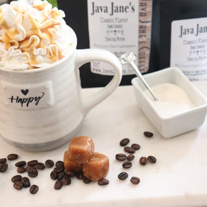 May include: A white ceramic mug with the word "Happy" and a heart, filled with a coffee drink topped with whipped cream and caramel drizzle. Caramel candies and coffee beans are scattered on a white surface.