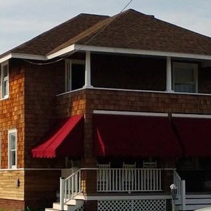 May include: A two-story house with a brown shingle exterior, white trim, and a dark brown roof. The house has a porch with red awnings and white railings. A car is parked in front of the house.