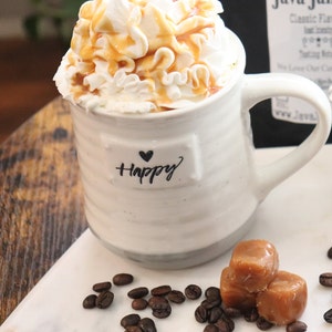 May include: A white ceramic mug filled with a whipped cream and caramel drink. The mug has the word "Happy" and a heart symbol printed on it. Coffee beans and caramel candies are scattered around the mug, suggesting a coffee shop theme.
