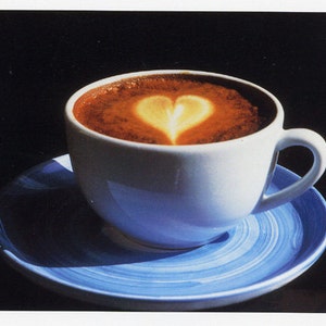 May include: A white ceramic coffee cup filled with a dark beverage, topped with a heart-shaped design in a lighter color. The cup sits on a blue saucer with a spiral pattern, against a dark background.