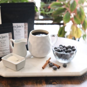 May include: A still life arrangement featuring coffee, blueberries, and cinnamon sticks. A white mug filled with coffee sits on a marble tray alongside a small creamer, sugar, and two bags of coffee beans. A glass bowl holds blueberries.
