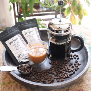 May include: A coffee setup featuring a French press, a double-walled glass of coffee, and two bags of Java Jane's coffee. Coffee beans and a measuring spoon are scattered on a dark wooden tray. The bags have the text "Classic Flavor" and "We Love Our Customers".