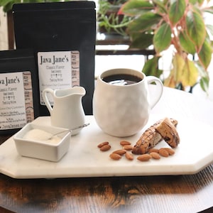 May include: A coffee setup featuring a white mug filled with dark coffee, biscotti, and almonds on a marble tray. Two black bags of coffee and a small creamer are also present. The coffee bags have the text "Java Jane's Classic Flavor".