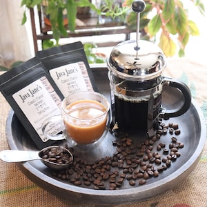 May include: A wooden tray holds a French press, a double-walled glass of coffee, and two black coffee bags labeled "Java Jane's." Coffee beans and a measuring spoon are scattered around the tray, creating a coffee-themed display.
