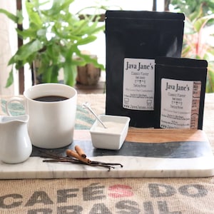 Puede incluir: Una composición de café con una taza blanca llena de café negro, una jarrita de crema y un pequeño cuenco de azúcar. Se ven dos bolsas de café Java Jane's, junto con vainas de vainilla y palitos de canela.