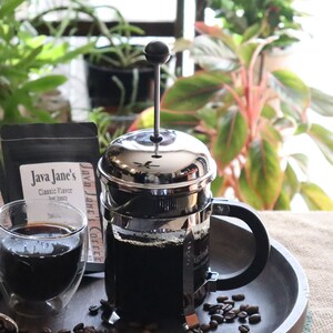 Puede incluir: Una cafetera francesa de acero inoxidable llena de café oscuro, junto a un vaso de café y una bolsa de granos de café Java Jane's Classic Flavor. La cafetera francesa está sobre una bandeja de madera con granos de café esparcidos.