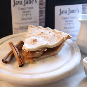 May include: A slice of apple pie dusted with powdered sugar on a white marble plate, with two cinnamon sticks. A white creamer and sugar bowl are also present. Two bags of coffee are in the background, with the text "Java Jane's".