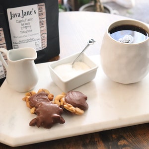 May include: A coffee setup featuring a white mug filled with coffee, a small creamer, a sugar bowl, and chocolate-covered cashews on a marble tray. Two bags of "Java Jane's" coffee are in the background.