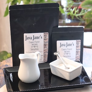 May include: A still life featuring two black coffee bags labeled "Java Jane's Classic Flavor", a small white creamer, and a white sugar bowl with a spoon, all arranged on a black tray. The bags have white labels with text.