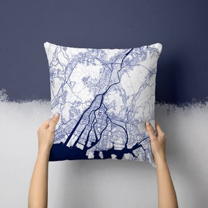Puede incluir: Cojín cuadrado blanco con un diseño de mapa azul detallado. El mapa presenta líneas intrincadas que representan carreteras, vías fluviales y áreas urbanas. El cojín tiene un borde azul marino sólido en la parte inferior. El cojín se sostiene contra una pared blanca y azul marino.