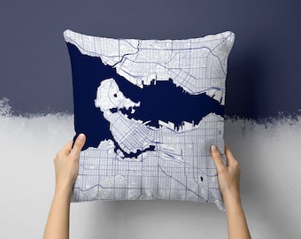 May include: A square pillow featuring a detailed map of a city in navy blue and white. The map design covers the entire surface of the pillow, showcasing streets, waterways, and urban areas. The pillow is held up against a two-tone blue wall.