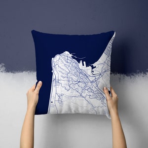 Puede incluir: Un cojín cuadrado con un fondo azul marino y un diseño de mapa detallado en blanco y azul. El mapa representa una ciudad costera con carreteras y edificios. El cojín está sostenido por dos manos.
