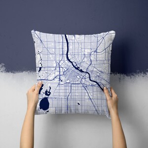 Puede incluir: Un cojín cuadrado con un diseño de mapa detallado en azul y blanco. El mapa muestra un plano de la ciudad con calles, vías fluviales y parques. El cojín se sostiene contra una pared azul oscuro.