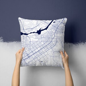 May include: A white square pillow with a detailed blue map design. The map features intricate street layouts, a river, and other geographical details. The pillow is held up by two hands against a two-tone blue and white background.