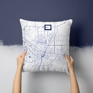 Pode incluir: Almofada quadrada branca com um design detalhado de mapa da cidade em azul. O mapa apresenta layouts de ruas intrincados e um marco proeminente destacado com um quadrado azul escuro. A almofada é segurada contra um fundo azul e branco de duas cores.