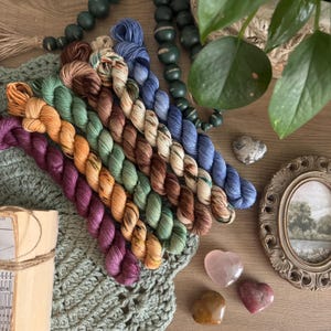 May include: A selection of multi-coloured yarn skeins in shades of purple, orange, green, and blue, arranged on a green crocheted piece. Decorative beads, heart-shaped stones, and a framed picture create a cosy, artistic display.