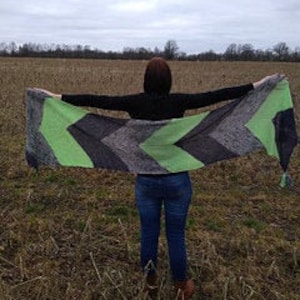 May include: A woman stands in a field, holding a large, rectangular shawl. The shawl features a chevron pattern in shades of green, gray, and black. The ends of the shawl have small, green tassels. The woman is wearing a black shirt and blue jeans.