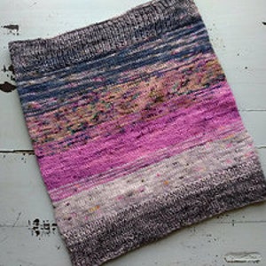 May include: A hand-knitted cowl in a gradient of colors, including shades of pink, purple, blue, brown, and gray. The cowl is made of yarn with a textured appearance. The knitted fabric is soft and appears to be warm.