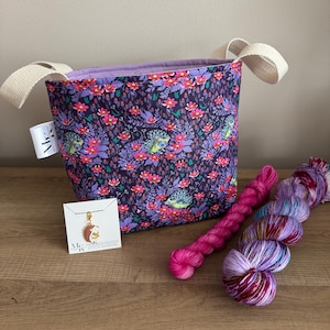 Może przedstawiać: Materiałowa torba na projekty dziewiarskie z kwiatowym i jeżowym wzorem, kremowymi uchwytami i fioletowym wnętrzem. Dwa motki przędzy w odcieniach różu i fioletu leżą obok torby, wraz z małą metką i wisiorkiem.