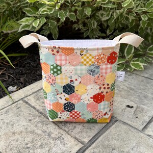 May include: A fabric storage basket featuring a patchwork design of colorful hexagons in various patterns including strawberries, gingham, and floral prints. The basket has two cream-colored fabric handles and a solid white interior lining.