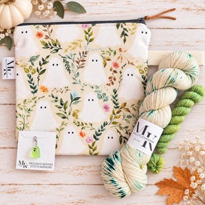 May include: A zippered pouch featuring a cream-colored fabric with a repeating pattern of white ghosts, green leaves, and small flowers. Beside it are two skeins of yarn in cream and green, and a small ghost-shaped stitch marker on a card.