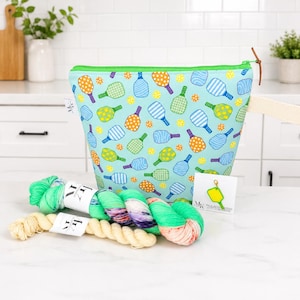 Può includere: Una pochette con cerniera azzurra con un motivo ripetuto di racchette da pickleball colorate e piccoli punti. Insieme alla pochette ci sono due matasse di filato nei toni del verde e della crema, oltre a un cartoncino con un segnapunti giallo.