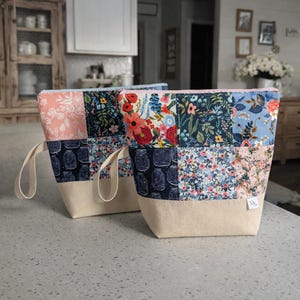May include: Two patchwork fabric tote bags with cream-coloured handles. The bags feature a variety of floral and jar patterns in pink, blue, and navy. The bottom of the bags is a solid cream colour.