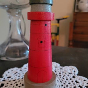 May include: A small wooden lighthouse figurine with a red body, a gray base, and a green roof. The lighthouse has a small window and a black dot on the side.