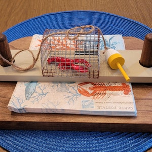 May include: A wooden napkin holder with a lobster trap design. The holder has two wooden posts on either side and a rope wrapped around the top. A red lobster is inside the trap. The holder is sitting on a blue and white napkin with a lobster and seaweed design. The text "CARTE POSTALE" is printed on the napkin.