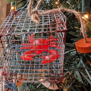 May include: A handmade Christmas ornament featuring a red plastic crab inside a small wire cage. The cage is trimmed with red paint and suspended by a twine loop. An orange disc is attached to the twine.