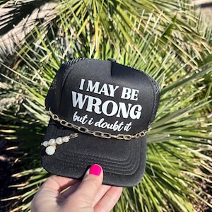 May include: Black trucker hat with the text "I MAY BE WRONG but I doubt it" in white. A gold chain and a pearl and rhinestone pin are attached to the hat. The hat is held up by a hand with bright pink nail polish.