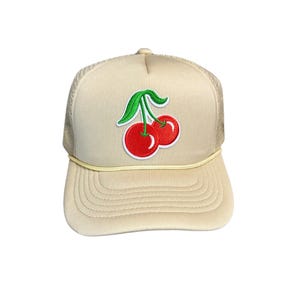 May include: A beige trucker hat with a red cherry embroidered on the front. The cherry has a green stem and leaves.