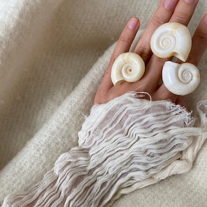 Könnte beinhalten: Eine Hand trägt drei weiße Muschelringe. Die Hand ruht auf einer weißen, flauschigen Oberfläche.