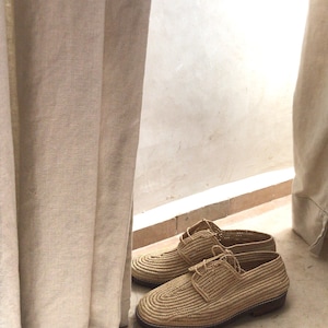 K&ouml;nnte beinhalten: Ein Paar geflochtene Strohschuhe mit Schn&uuml;rsenkeln. Die Schuhe sind hellbraun und haben eine leicht spitz zulaufende Spitze. Die Schuhe stehen auf einem hellbraunen Fliesenboden.