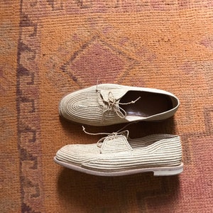 K&ouml;nnte beinhalten: Ein Paar beige gewebte Schuhe mit wei&szlig;en Sohlen. Die Schuhe befinden sich auf einem braunen Teppich mit einem gemusterten Design.