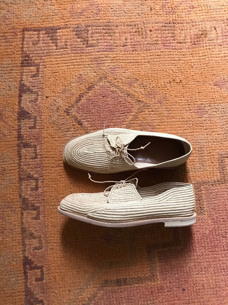 K&ouml;nnte beinhalten: Ein Paar beige gewebte Schuhe mit wei&szlig;en Sohlen. Die Schuhe befinden sich auf einem braunen Teppich mit einem gemusterten Design.