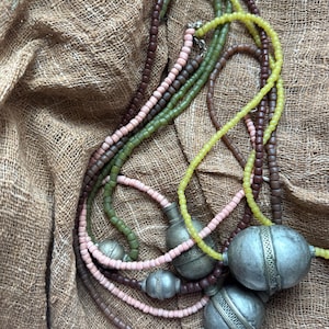 Puede incluir: Una colección de collares de cuentas en varios colores, incluyendo marrón, verde, rosa y amarillo. Los collares presentan pequeñas cuentas cuadradas y colgantes de metal plateado con diseños intrincados. Se exhiben sobre una tela marrón texturizada.