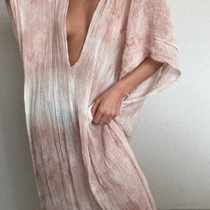 May include: A flowing, blush-colored dress with a deep V-neck. The dress has a loose, draped design and short sleeves. The fabric appears lightweight and textured, with subtle variations in color. A beaded necklace is visible at the neckline.