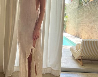 May include: A sleeveless, floor-length, cream-colored dress with high side slits. The dress has a textured, semi-sheer fabric and a high neckline. The background includes a window with a view of a pool and a striped armchair.
