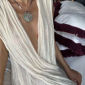 May include: A woman wearing a white, sheer, flowing dress with a silver heart-shaped pendant necklace.
