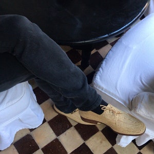 K&ouml;nnte beinhalten: Ein Paar beige gewebte Schuhe mit schwarzen Schn&uuml;rsenkeln auf einem schwarz-wei&szlig;en Schachbrettmuster-Fliesenboden. Die Schuhe werden von einer Person getragen, die eine dunkelblaue Jeans tr&auml;gt.