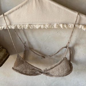 Puede incluir: Top de bikini tejido beige con tirantes ajustables. La parte superior está hecha de un material tejido texturizado y presenta un detalle de nudo retorcido en el centro. El fondo es una pared de color claro con una superficie texturizada.