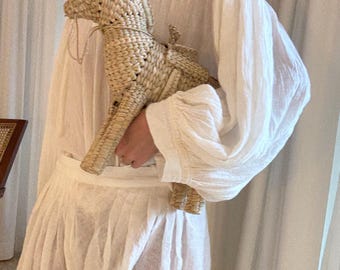 Può includere: Una statuetta di cavallo in paglia intrecciata con criniera, coda e briglia dettagliate. Il cavallo è di colore marrone chiaro e ha una superficie strutturata. La statuetta è tenuta contro un abito bianco.