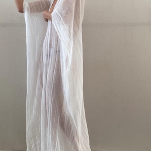 May include: A floor-length white kaftan made of a lightweight, semi-sheer, textured fabric. The garment features wide, flowing sleeves, a deep V-neckline, and a loose, relaxed silhouette that drapes down to the floor, creating a breezy and airy appearance.