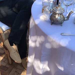 K&ouml;nnte beinhalten: Eine wei&szlig;e Tischdecke mit einer silbernen Teekanne, einem Glas Tee und einem silbernen L&ouml;ffel. Eine Person in blauen Jeans und braunen Schuhen sitzt an einem Tisch mit einer wei&szlig;en Tischdecke.