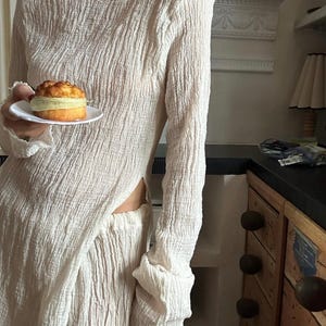 May include: A white, long-sleeved, loose-fitting dress with a side slit. The dress is made of a textured fabric and has a high neckline. The dress is being worn by a person holding a small pastry.