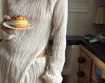 Puede incluir: Un vestido blanco de manga larga y holgado con una abertura lateral. El vestido está hecho de una tela texturizada y tiene un cuello alto. El vestido lo lleva puesto una persona que sostiene un pequeño pastel.