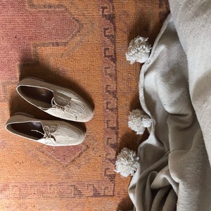 K&ouml;nnte beinhalten: Zwei Paar beige gewebte Schuhe mit Schn&uuml;rsenkeln auf einem braunen Teppich mit einer wei&szlig;en Decke mit Bommeln an der Seite.