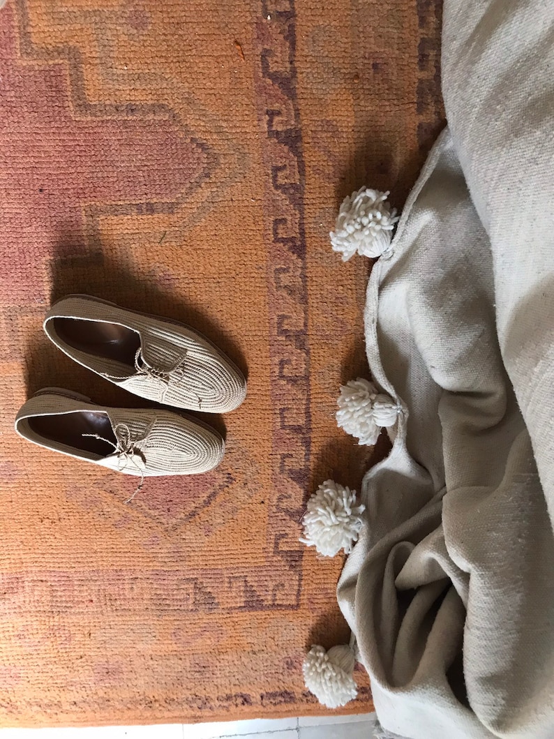 K&ouml;nnte beinhalten: Zwei Paar beige gewebte Schuhe mit Schn&uuml;rsenkeln auf einem braunen Teppich mit einer wei&szlig;en Decke mit Bommeln an der Seite.